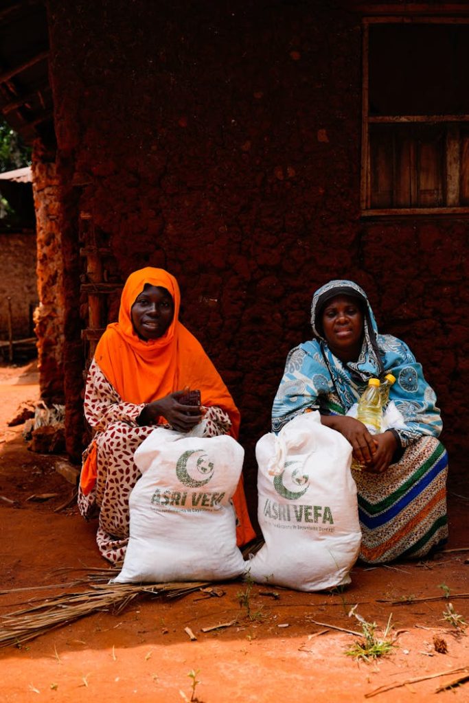 Two women sit by a mud house with relief bags, showing community aid.